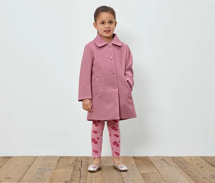 Mädchen steht vor einer weißen Wand und trägt einen rosa Mantel, Leggings mit Sternen und silberne Schuhe.