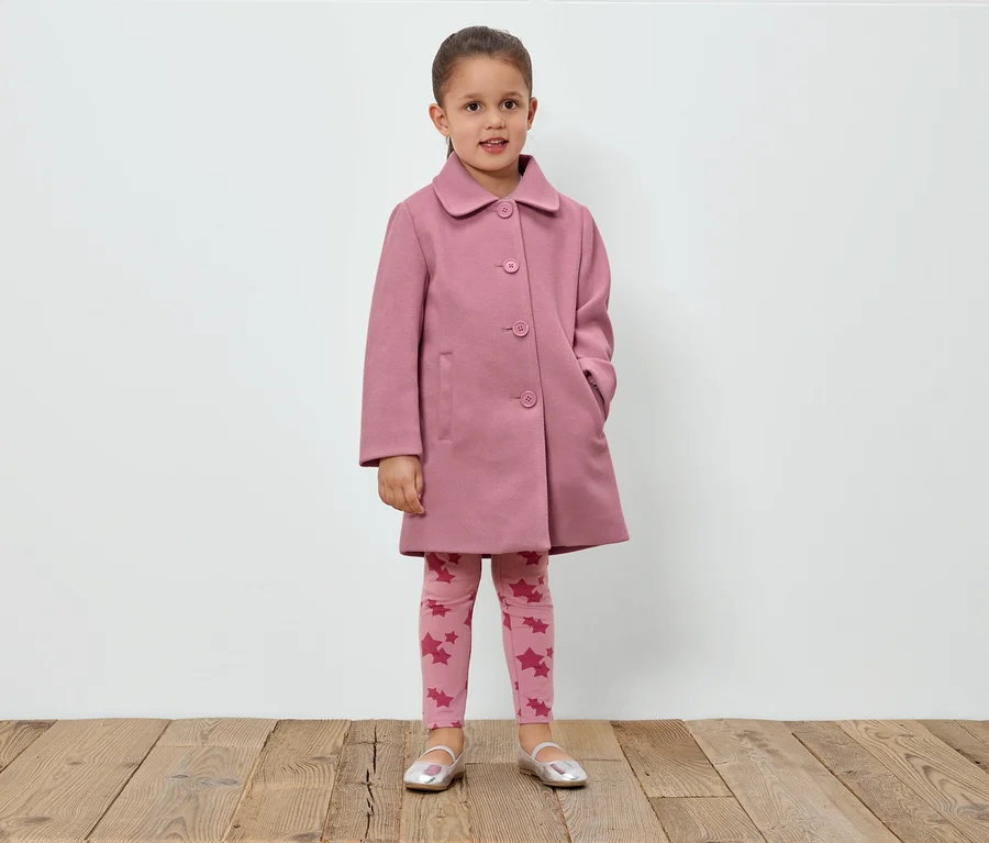 Mädchen steht vor einer weißen Wand und trägt einen rosa Mantel, Leggings mit Sternen und silberne Schuhe.