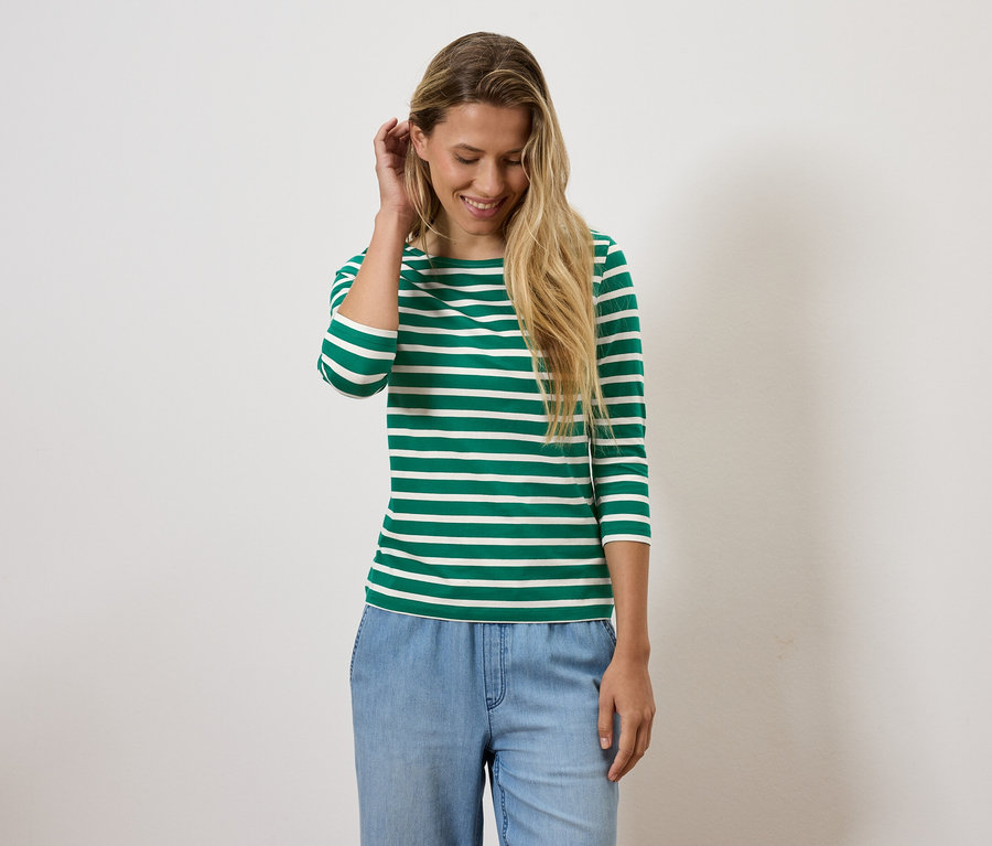 Blonde Frau lächelt und richtet ihr Haar, trägt ein grün-weiß gestreiftes Shirt und blaue Jeans.