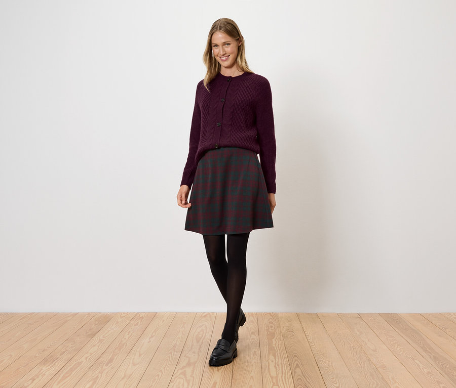 Eine Frau in einem burgunderroten Pullover, einem dunklen Rock, einer schwarzen Strumpfhose und Schuhen steht auf einem Holzboden.