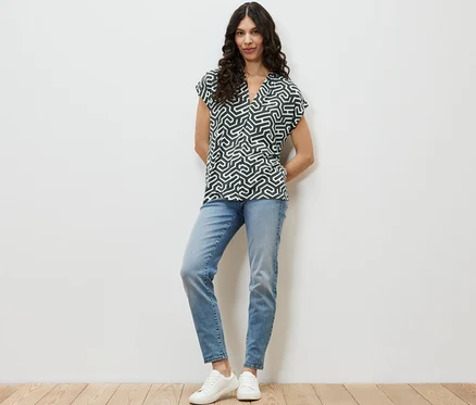 Frau steht und trägt ein grünes Blusenshirt mit V-Ausschnitt, blaue Jeans und weiße Turnschuhe.