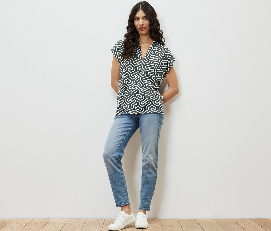 Frau steht und trägt ein grünes Blusenshirt mit V-Ausschnitt, blaue Jeans und weiße Turnschuhe.