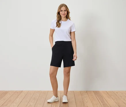 Frau mit weißem T-Shirt und schwarzen Shorts steht vor einer Wand.