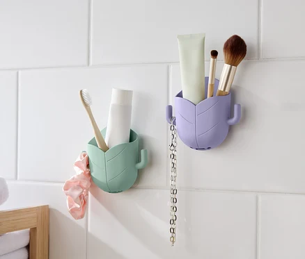 Zwei blattförmige Behälter hängen an einer weißen, gefliesten Wand. Der linke Behälter ist mintgrün und enthält eine Zahnbürste und eine Tube. Ein rosa Scrunchie hängt daneben. Der rechte Behälter ist lavendelfarben und enthält eine Tube, einen Pinsel und eine Kette.