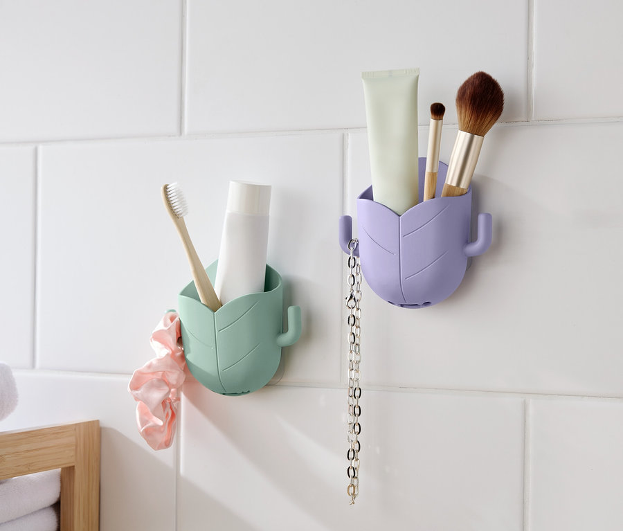 Zwei blattförmige Behälter hängen an einer weißen, gefliesten Wand. Der linke Behälter ist mintgrün und enthält eine Zahnbürste und eine Tube. Ein rosa Scrunchie hängt daneben. Der rechte Behälter ist lavendelfarben und enthält eine Tube, einen Pinsel und eine Kette.