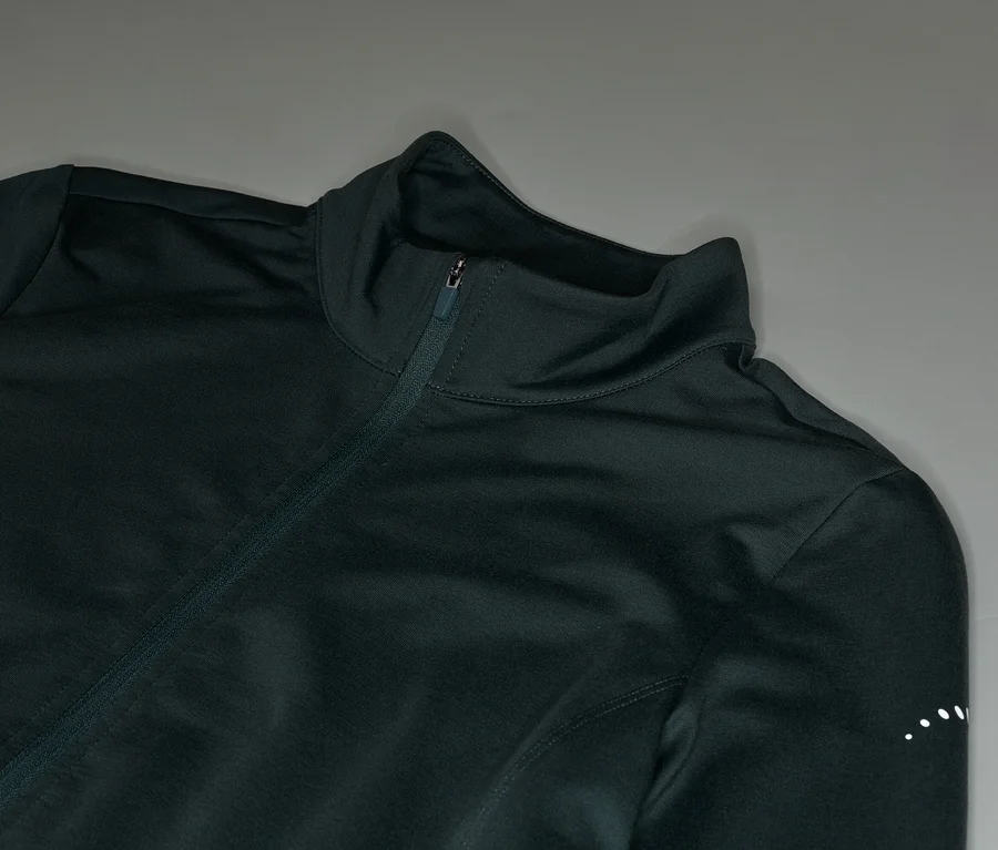 Nahaufnahme einer dunkelgrünen Trainingsjacke mit Reißverschluss und Stehkragen vor grauem Hintergrund.