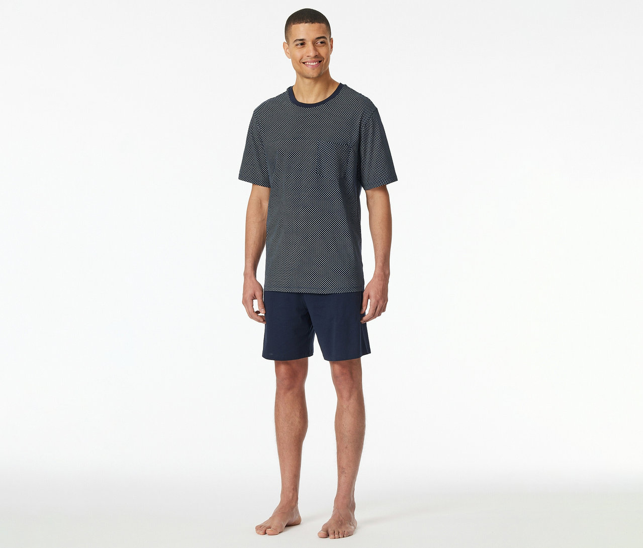 Mann steht barfuß in SCHIESSER Shorty-Pyjama-Set aus marineblauem, kurzärmligem Punkt-Shirt und Shorts vor weißem Hintergrund.