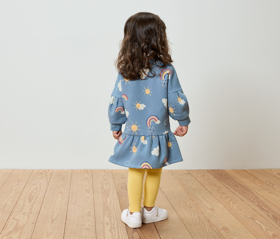 Ein kleines Mädchen steht mit dem Rücken zur Kamera und trägt ein blaues Kleid mit einem Muster aus Sonne, Wolken und Regenbogen, gelbe Leggings und weiße Schuhe.