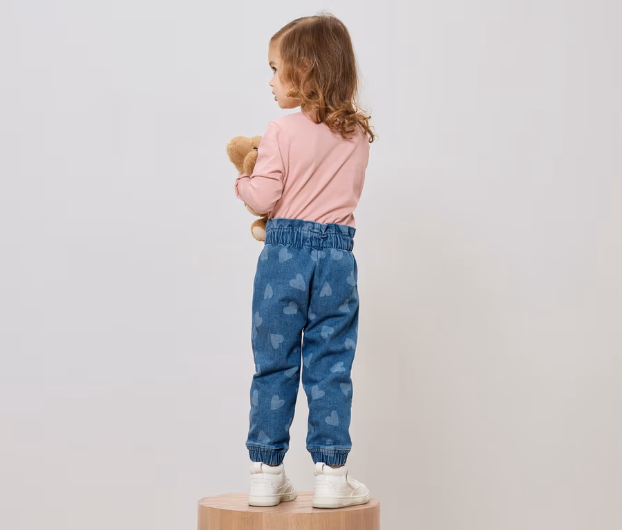 Ein kleines Mädchen mit lockigem Haar steht mit dem Rücken zur Kamera und umarmt einen Teddybären. Sie trägt ein rosafarbenes Langarmshirt, blaue Jeans mit Herzmuster und weiße Turnschuhe.