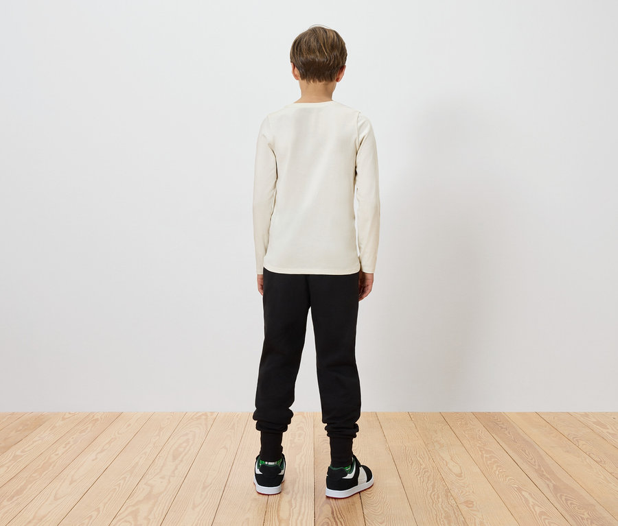 Ein Kind steht mit dem Rücken zum Betrachter, trägt ein beiges Langarmshirt, schwarze Hosen und grüne und schwarze Turnschuhe.