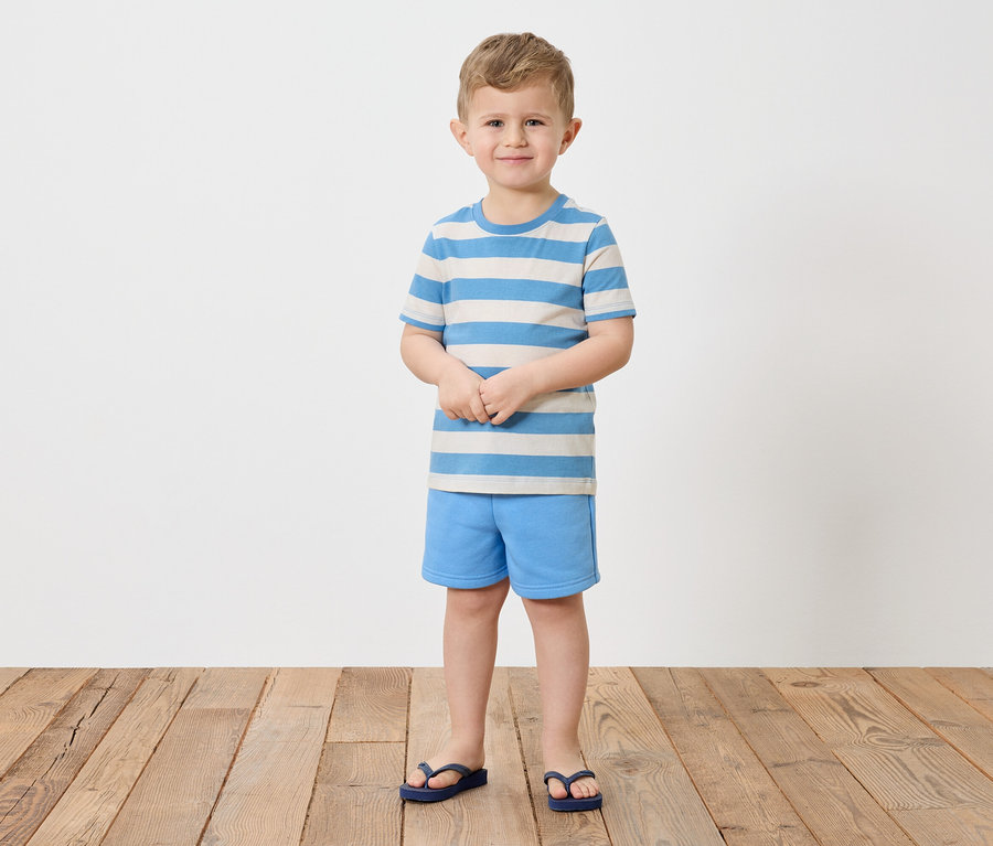 Ein Junge mit blonden Haaren steht barfuß auf einem Holzboden und trägt ein gestreiftes blau-weißes T-Shirt, blaue Shorts und blaue Flip-Flops.
