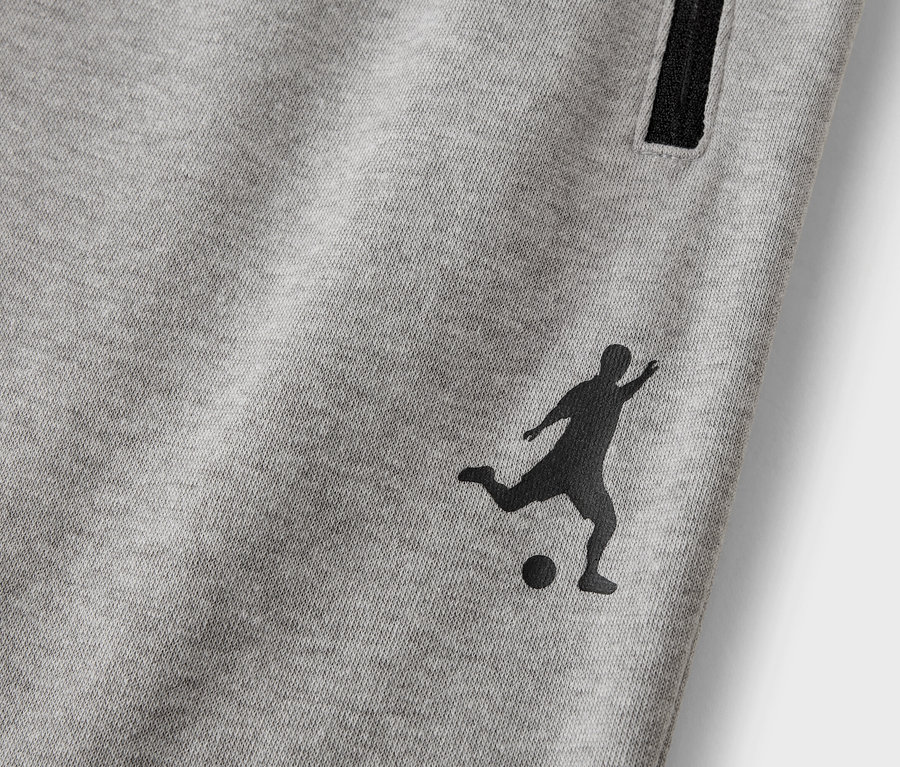 Nahaufnahme einer grauen Trainingshose mit einer schwarzen Silhouette eines Fußballspielers und einer Reißverschlusstasche.