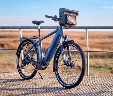 Blaues E-Bike mit FISCHER Lenkertasche PLUS+ Terra steht auf einem Holzsteg vor einer Dünenlandschaft.