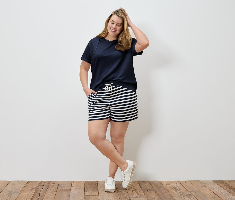 Frau in marineblauem T-Shirt und gestreiften Shorts posiert vor einer Wand. Sie hat eine Hand im Haar und eine in der Hosentasche.