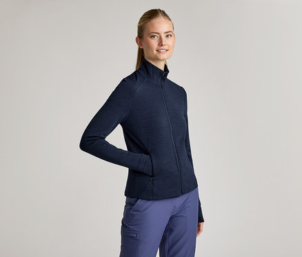 Frau mit Zopf trägt eine marineblaue Jacke mit Reißverschluss und marineblaue Hose. Eine Hand steckt in der Jackentasche.