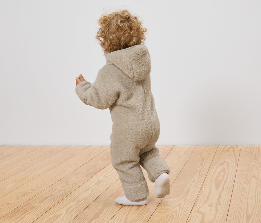 Ein Kind mit lockigem Haar trägt einen Baby-Teddy-Overall und weiße Socken, während es auf einem Holzboden vor einer weißen Wand steht.