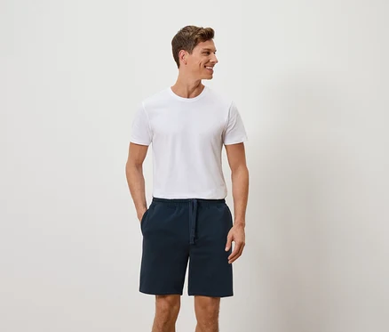 Mann mit weissem T-Shirt und blauen Shorts, die Hände in den Hosentaschen, lächelt zur Seite.