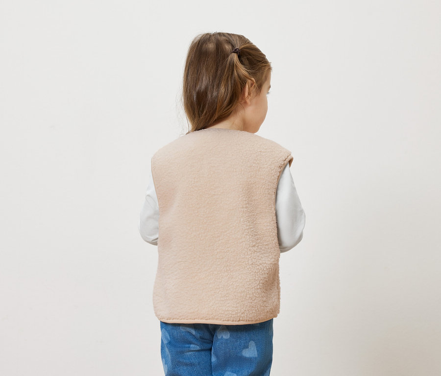 Rückansicht eines kleinen Mädchens mit Zopf, das eine beigefarbene Fleeceweste über einem weißen Langarmshirt und blauen Jeans mit Herzmuster trägt.