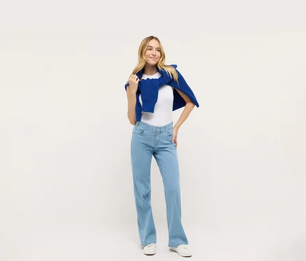 Frau posiert in einem weißen T-Shirt und blauen Angels Denim »Liz« Wide Leg Jeans, mit einem blauen Pullover über den Schultern.