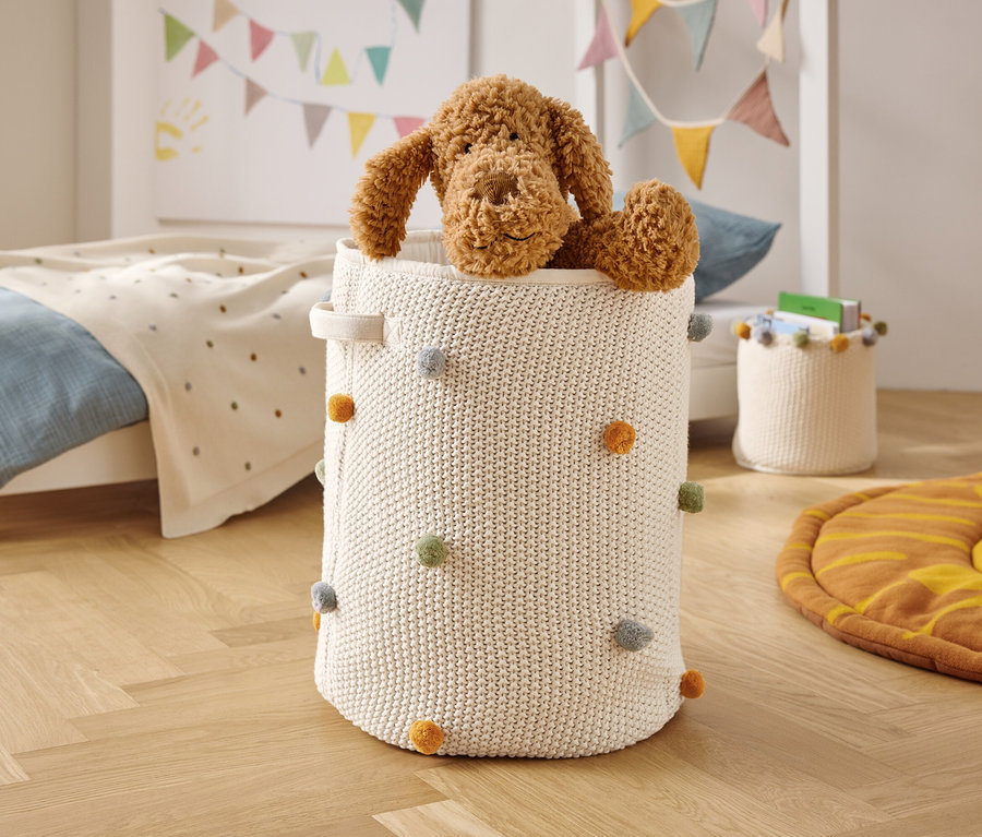 Ein brauner Stoffhund sitzt in einem weißen, gestrickten Korb mit bunten Bommeln.