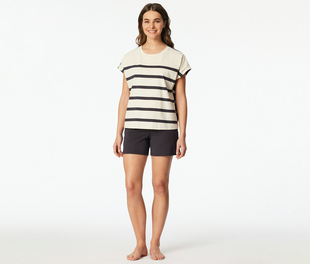 Lächelnde Frau trägt ein SCHIESSER Shorty-Set: ein cremefarbenes, gestreiftes Kurzarm-Oberteil und dunkelgraue Shorts. Sie ist barfuß.