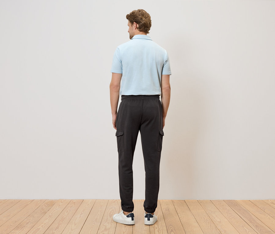 Mann von hinten in hellblauem Poloshirt und dunkler Cargohose vor weißer Wand und Holzboden.