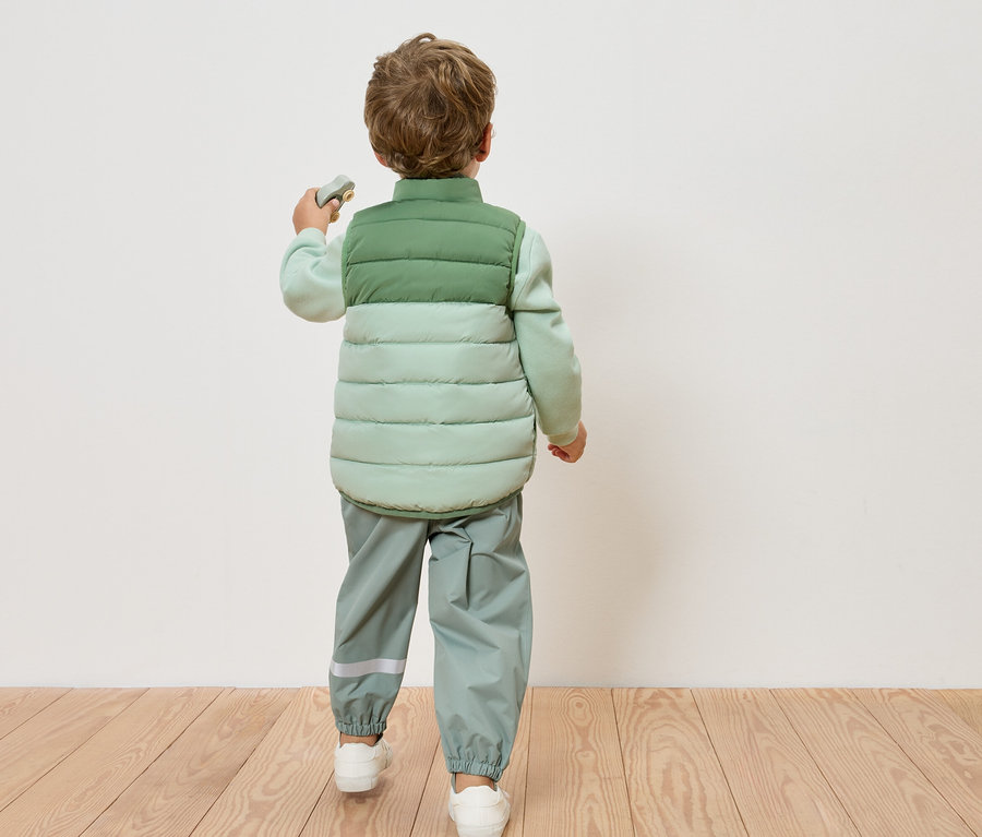 Ein Kind von hinten, trägt eine hellgrüne Steppweste und eine hellgrüne Hose, steht auf einem Holzboden vor einer weißen Wand.