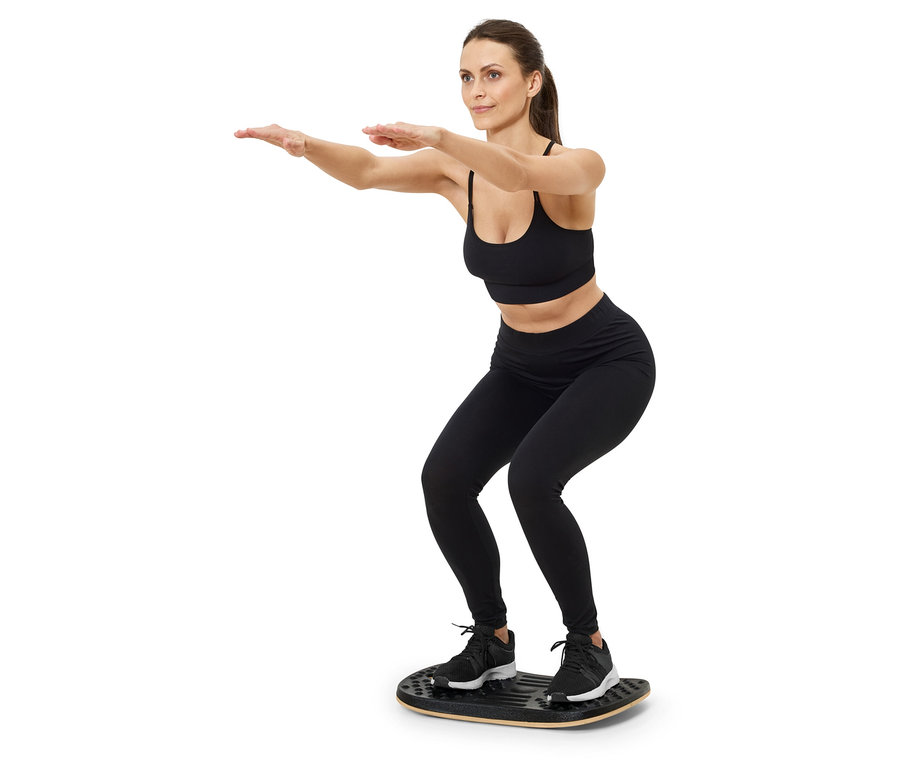 Frau in schwarzem Sport-BH und Leggings macht Kniebeugen auf einem Balance Board.