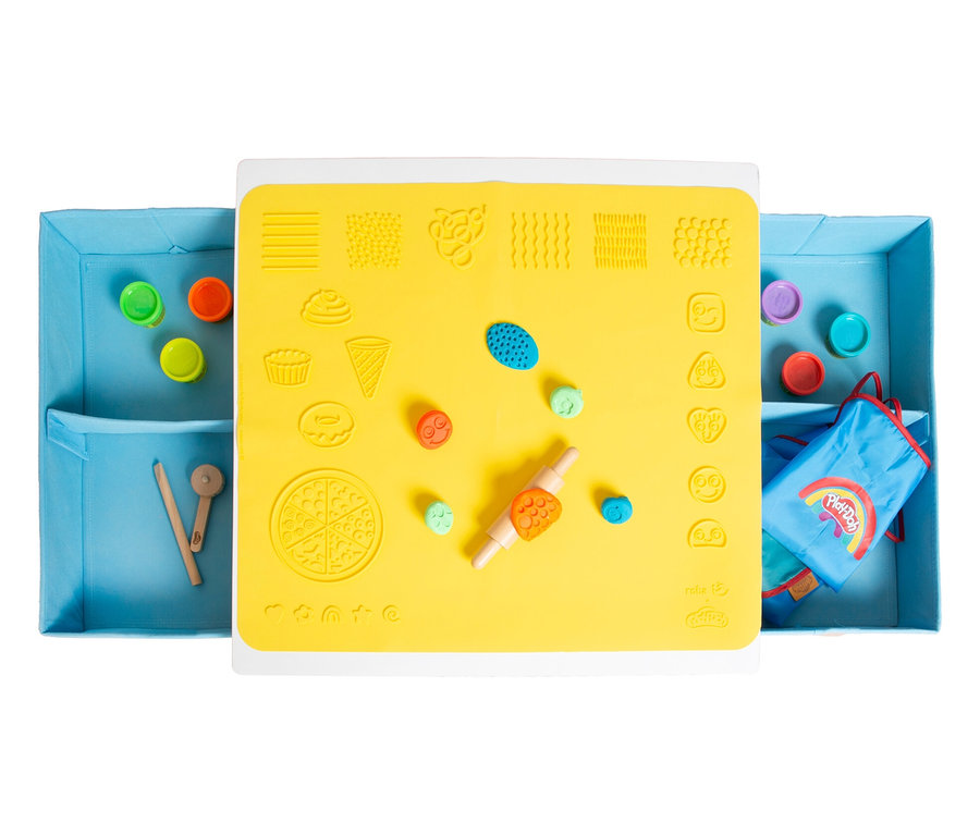 Draufsicht auf einen Tisch mit gelber Matte, Knete, Nudelholz und Förmchen. An den Seiten blaue Behälter mit Knete und Werkzeugen.