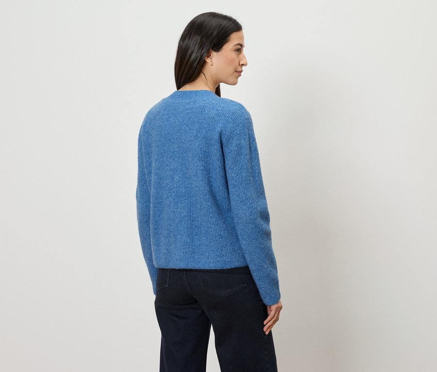 Eine Frau mit dunklen Haaren trägt einen blauen Pullover und dunkle Jeans, während sie vor einer weißen Wand steht.