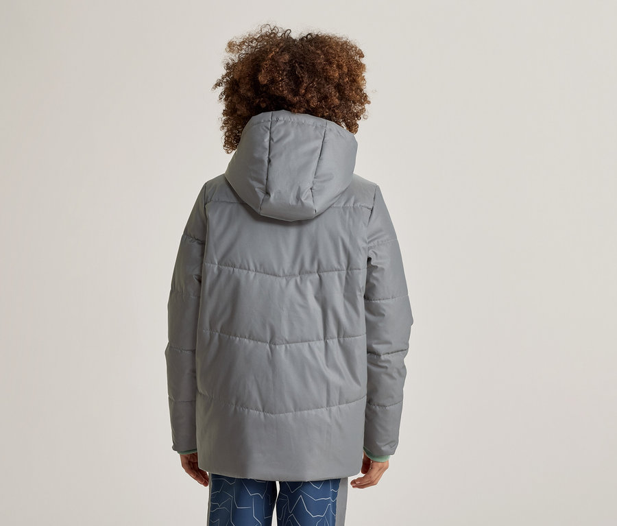 Person mit lockigem Haar von hinten, trägt eine graue, gesteppte Jacke mit Kapuze und blaue Hosen mit Muster.