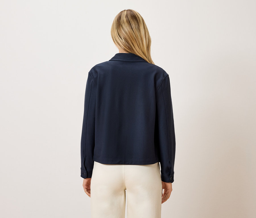 Rückansicht einer Frau mit blonden Haaren, die eine dunkelblaue Jacke und cremefarbene Hosen trägt.