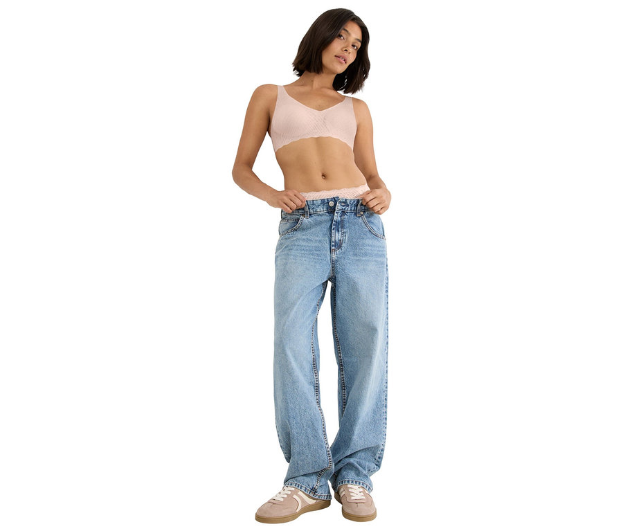 Frau mit kurzem braunem Haar, die einen hellrosa Spitzen-BH, eine weite, hellblaue Jeans und hellbraune Turnschuhe trägt.