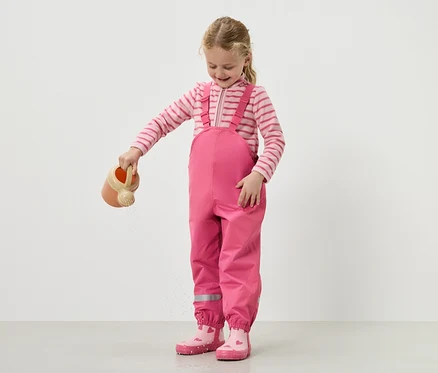 Ein Mädchen gießt Wasser mit einer Gießkanne, während sie eine rosa Latzhose und Stiefel trägt.