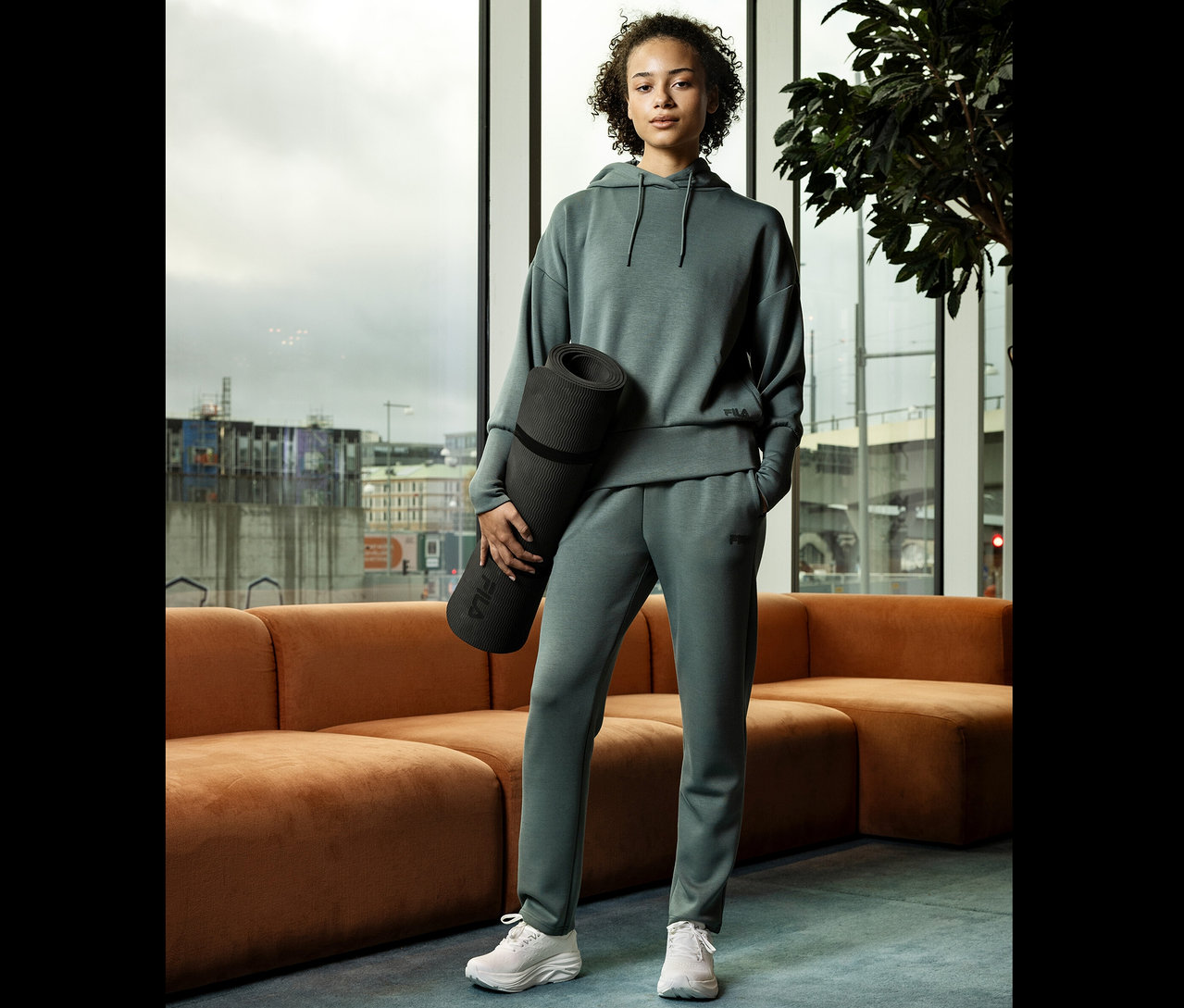 Junge Frau in grauer Trainingsbekleidung hält eine zusammengerollte Yogamatte vor einem Fenster.