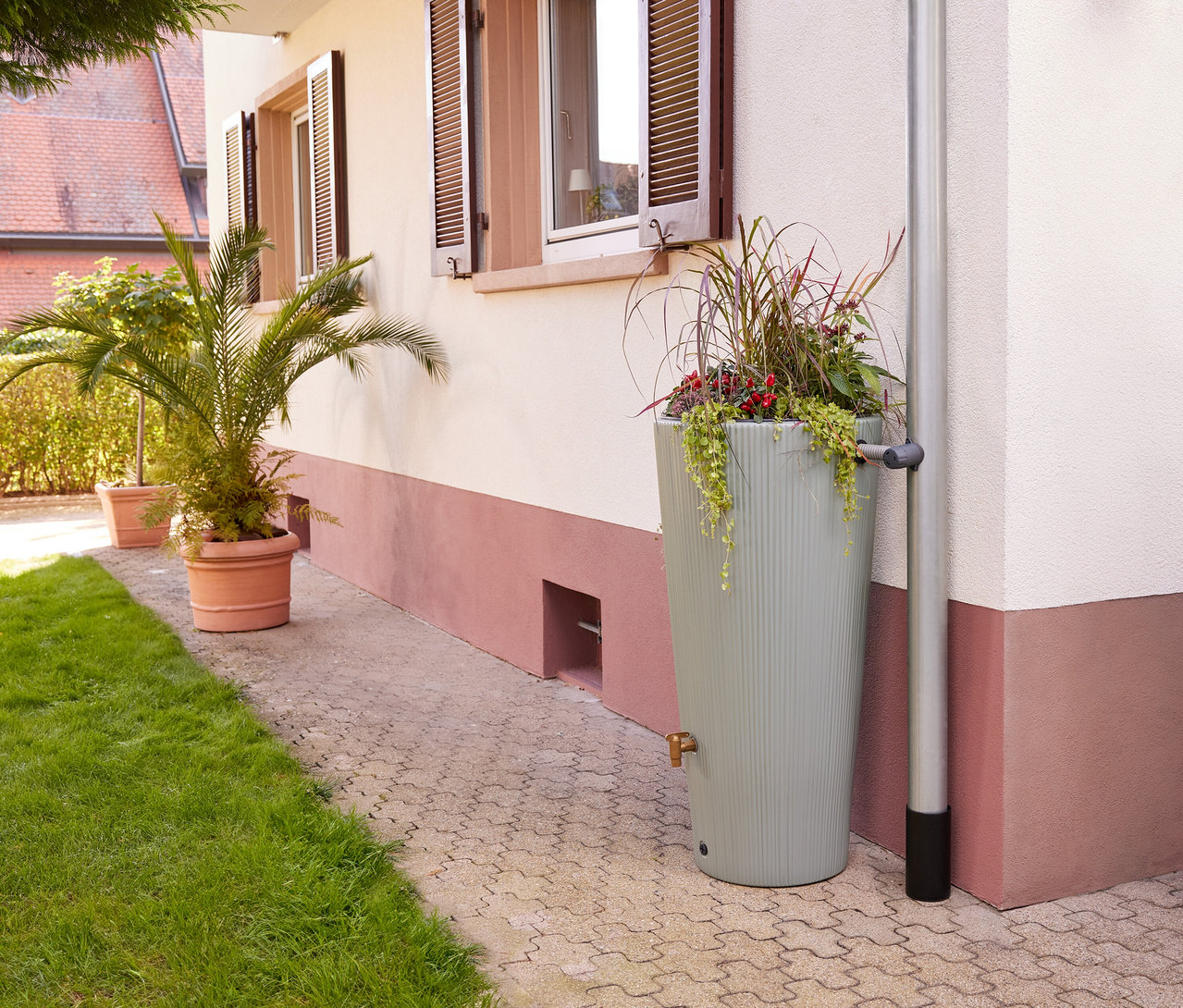 Vor einem Haus steht ein Garantia »Vaso« 2-in-1-Regenspeicher-Set mit Pflanzen befüllt neben einem Fallrohr.