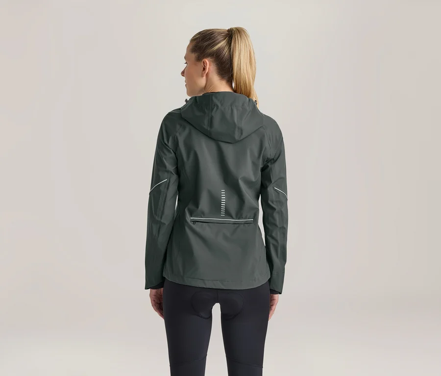Frau von hinten, trägt eine dunkelgrüne Kapuzenjacke mit reflektierenden Details am Rücken und an den Ärmeln sowie schwarze Radlerhosen. Ihr blondes Haar ist zu einem Pferdeschwanz gebunden.