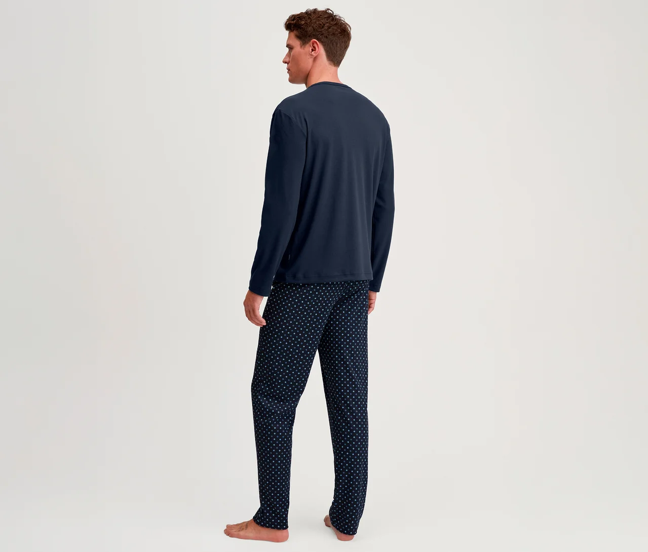 Mann von hinten, trägt ein dunkelblaues Langarmshirt und eine dunkelblaue Pyjamahose mit weißen Punkten, barfuß stehend.