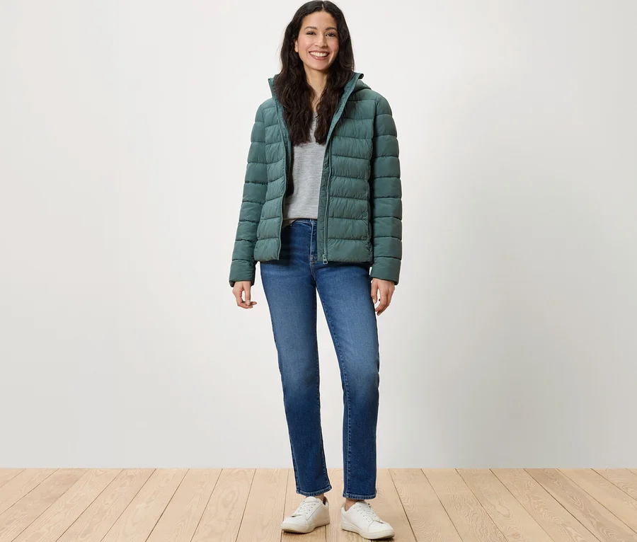 Eine Frau steht und lächelt in einer grünen Daunenjacke, blauen Jeans und weißen Turnschuhen.
