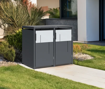Ein grauer Mülltonnencontainer mit zwei Türen steht auf einem gepflasterten Weg neben einem Haus.