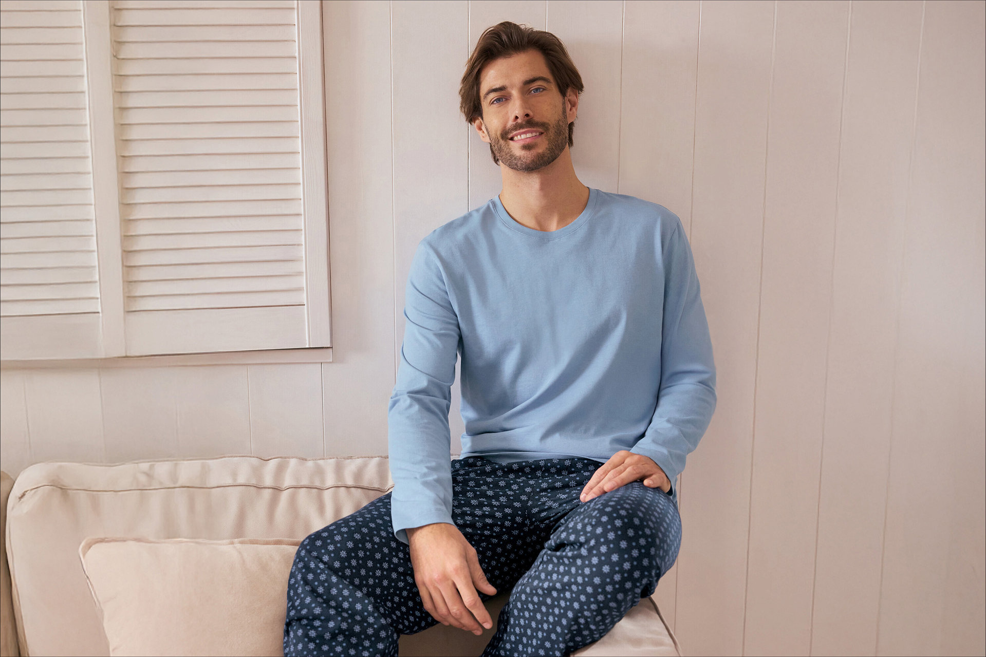 Ein lächelnder Mann mit Bart sitzt auf einem hellen Sofa und trägt ein hellblaues Langarmshirt und eine dunkelblaue Pyjamahose mit Muster. Im Hintergrund sind weiße Fensterläden zu sehen.