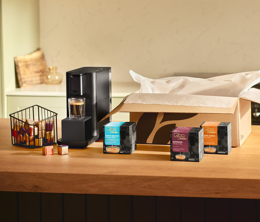 Schwarze Qbo Iconic, Dark Chrome Kaffeemaschine mit einem Espresso im Glas auf einer Holztheke. Daneben ein Korb mit bunten Qbo Caffè MINAS VERDE Kapseln, zwei lose Kapseln und drei Packungen: Qbo Caffè Grande KIRINYAGA BAYA, Qbo Espresso APANECA ANA und Caffè Crema, vor einem offenen Karton.