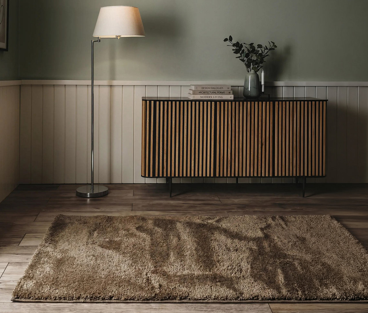 Ein gemütlicher Raum mit einem hellbraunen Hochflorteppich auf Holzboden. Links steht eine Stehlampe mit cremefarbenem Schirm, die ein Holz-Sideboard beleuchtet. Darauf stehen eine Vase mit Grünzeug, zwei Kerzen und Bücher. Die Wand ist oben salbeigrün, unten weiß getäfelt.