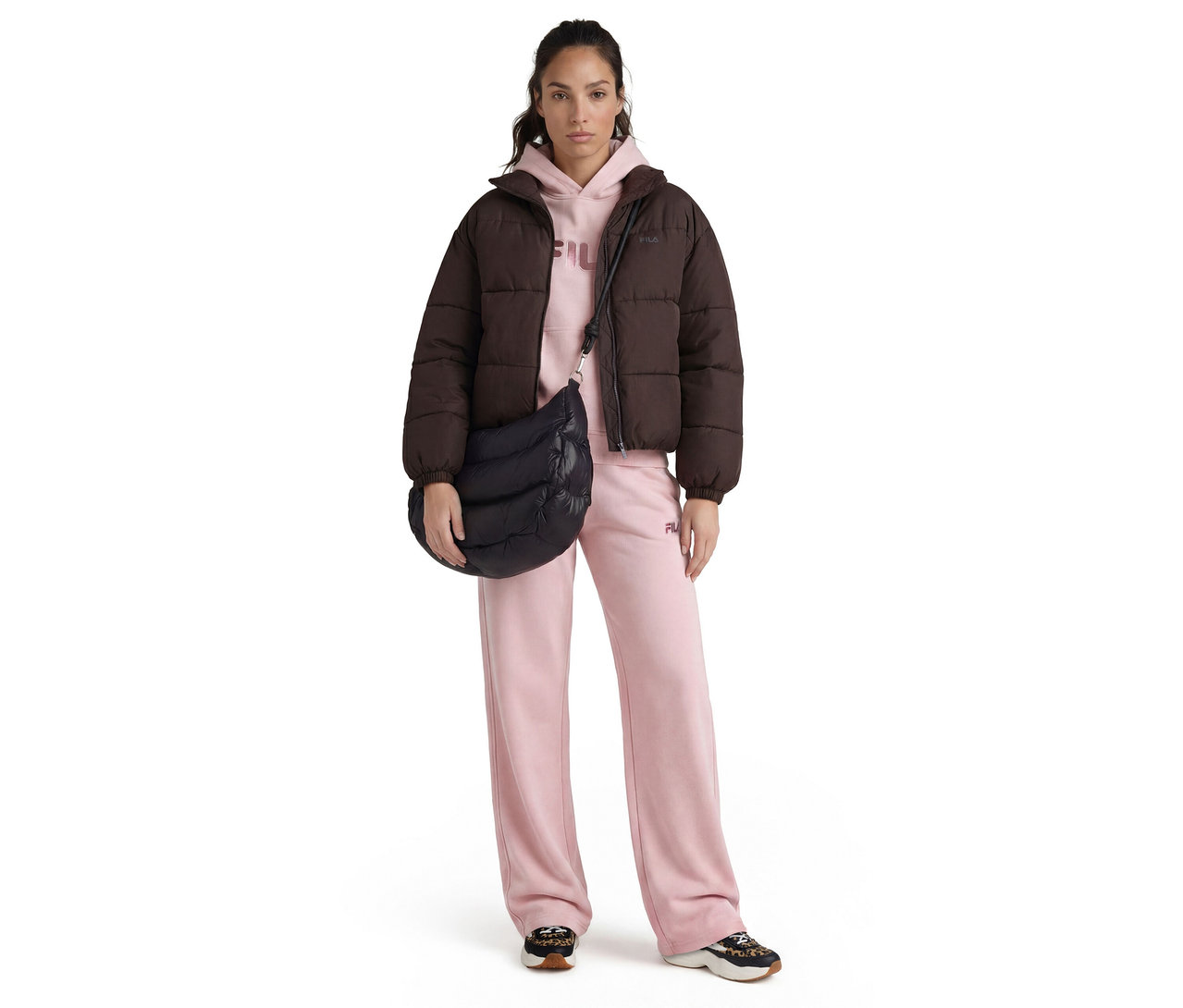 Eine Frau steht frontal, trägt eine dunkelbraune Pufferjacke, einen hellrosa Hoodie, hellrosa weite Jogginghosen und schwarz-leopard gemusterte Sneaker. Sie trägt eine schwarze gesteppte Umhängetasche.