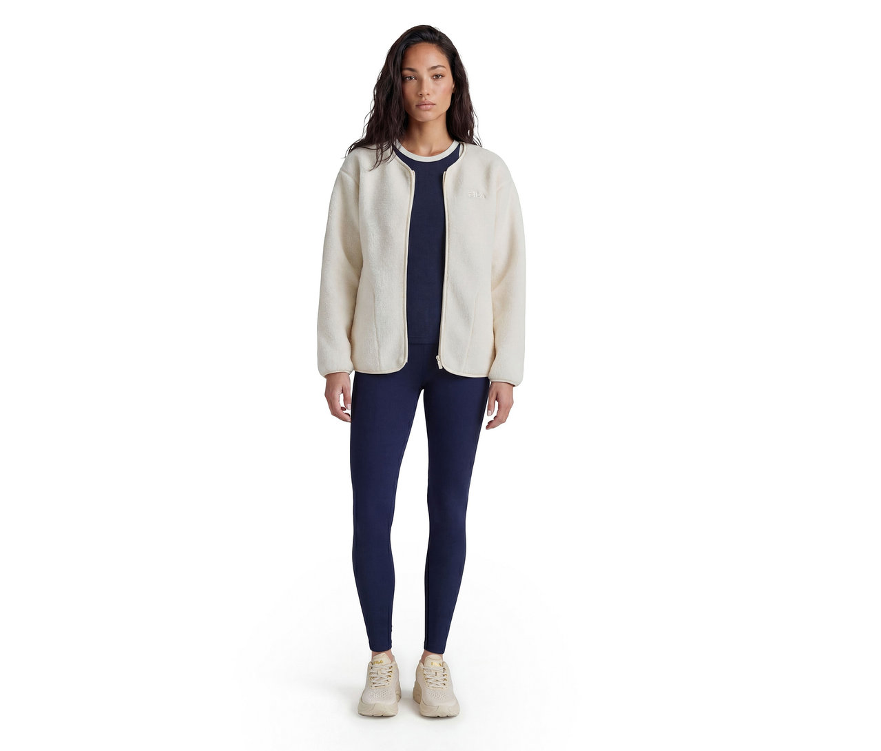 Frau steht, trägt eine cremefarbene Fleecejacke über einem marineblauen Oberteil und marineblauen Leggings, dazu helle Sneaker.