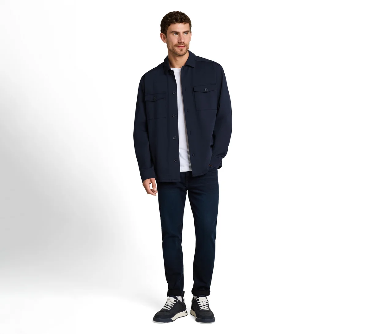 Mann in dunkelblauer Overshirt-Jacke, weißem T-Shirt, dunklen Jeans und dunklen Sneakers, der nach rechts blickt.