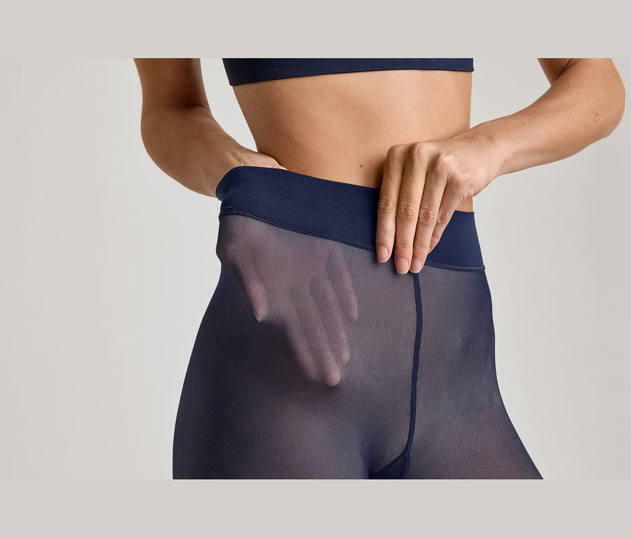 Nahaufnahme einer Frau in marineblauen, transparenten Strumpfhosen und passendem Sport-BH. Ihre Hände ziehen am Bund der Strumpfhose, wobei eine Hand durch den transparenten Stoff an ihrer Hüfte sichtbar ist.