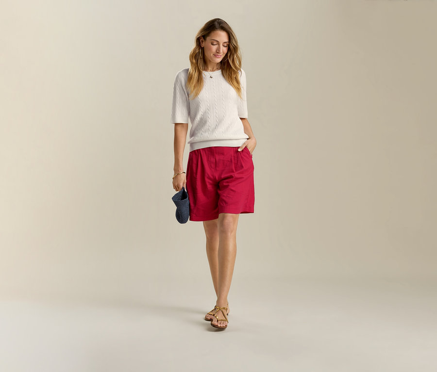 Frau in weißem Strickpullover und roten Leinen-Bermuda-Shorts, die eine Hand in der Tasche hat und ein blaues Visier hält.