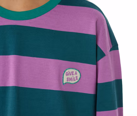 Nahaufnahme eines Kindes in einem SCHIESSER Kinder-Pyjama. Das Oberteil hat lila und dunkelgrüne Querstreifen, einen grünen Kragen und eine aufgestickte Sprechblase mit "GIVE A SMILE" auf der Brust.
