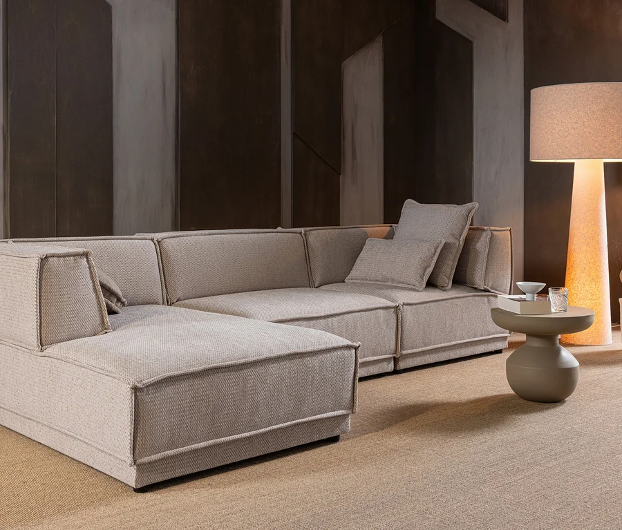 Ein modernes, beiges L-förmiges Sofa mit Zierkissen steht auf einem beigen Teppich. Daneben eine hohe Stehlampe mit warmem Licht, ein runder Beistelltisch mit Buch und Glas sowie ein kleiner Hocker mit Vase.